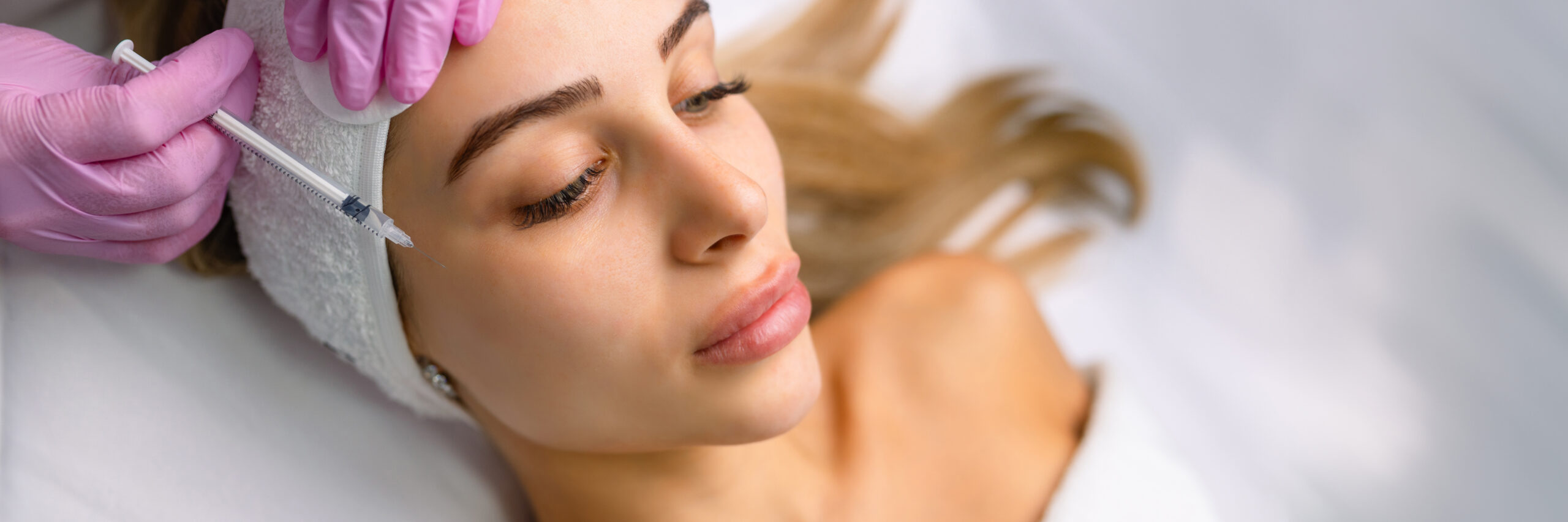 Close up of hands of young cosmetologist injecting botox in female face. She is standing and smiling. The woman is closed her eyes with relaxation.